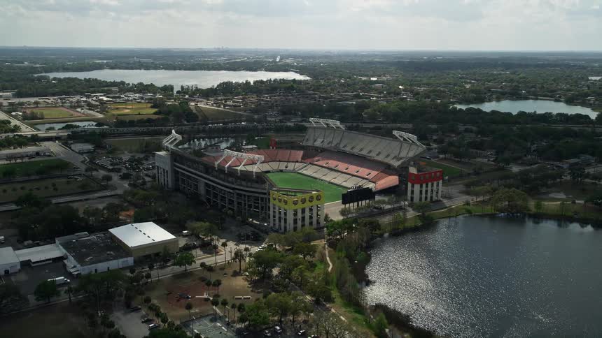 Approaching aerial view of Citrus Bowl football stadium and Lake Lorna Doone surrounded by greenery in downtown Orlando Florida