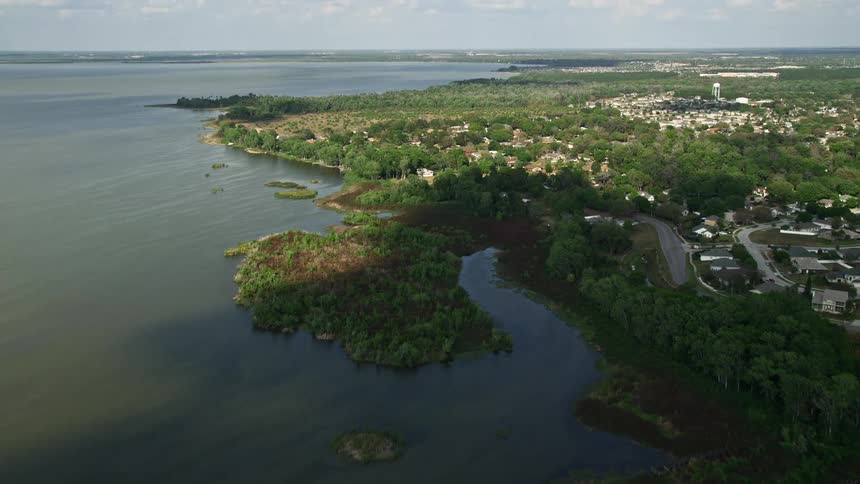 Approaching lakefront residences in Central Florida from a aerial view with a tilt up from the shoreline to reveal the area amidst partly cloudy skies and the shimmering waters of Lake Apopka