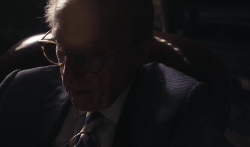 Close up of the face of a bespectacled elderly man sitting on a dimly lit room