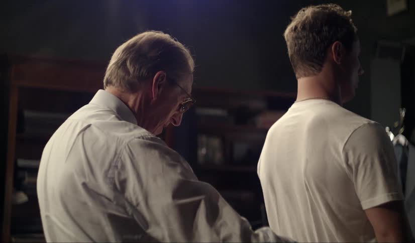 A tailor measuring the shoulders and backside of a man standing in front of him wearing a white shirt