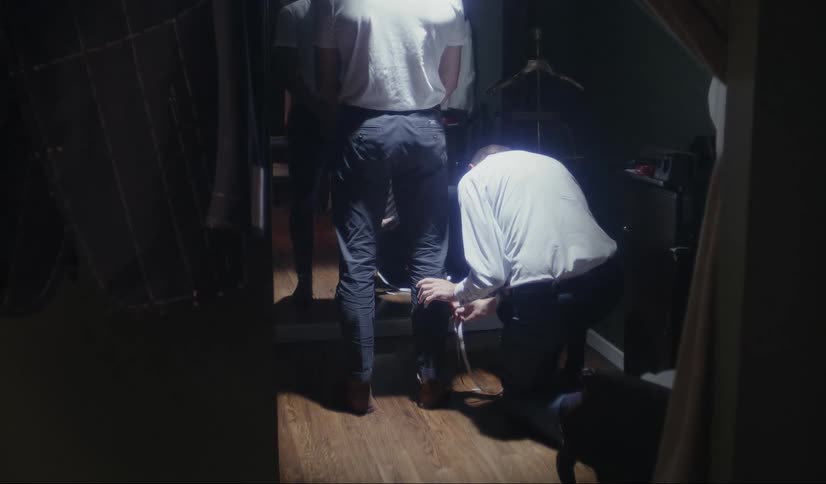 Tailor kneels to measure the lego of a client in a dimly lit dressing room
