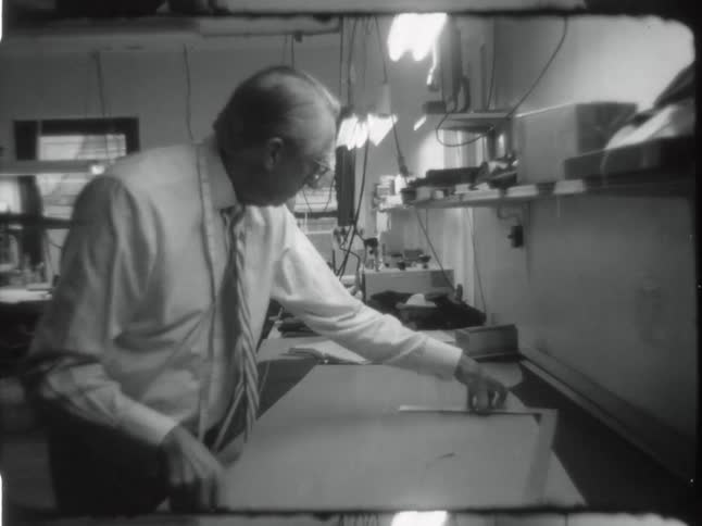 Black and white footage of a man measuring a sewing pattern caught on vintage film