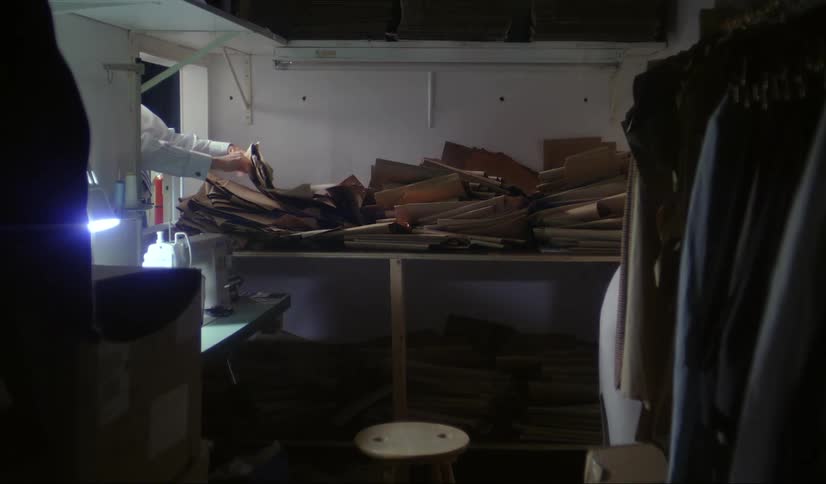 A small room with jackets hanging a small stool in front of a sewing machine and a persons hands stifling through a pile of brown papers
