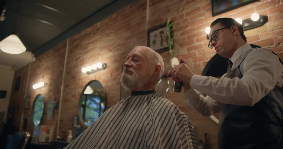 A barber combing and spraying a customers hair sitting in the chair in front of him