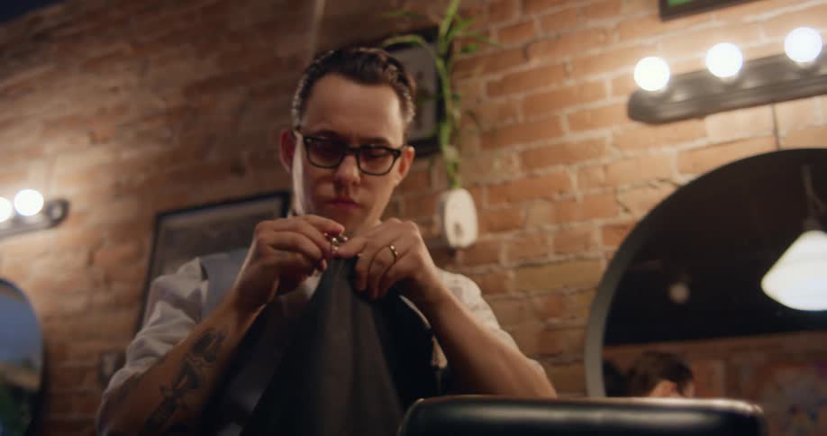 A barber stands in a shop while putting on a black apron and looking at the mirror