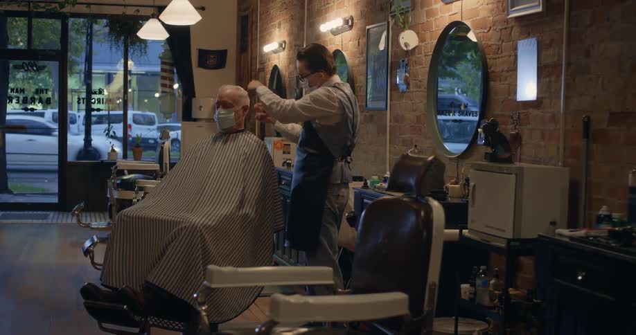 A young barber combs and trims an older mans hair in a shop at night