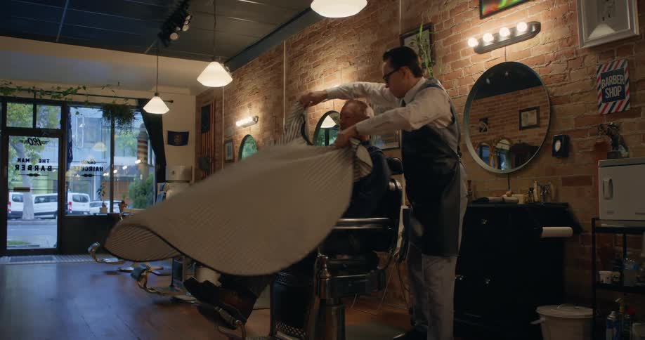 A young barber drapes a cloth over his older customer