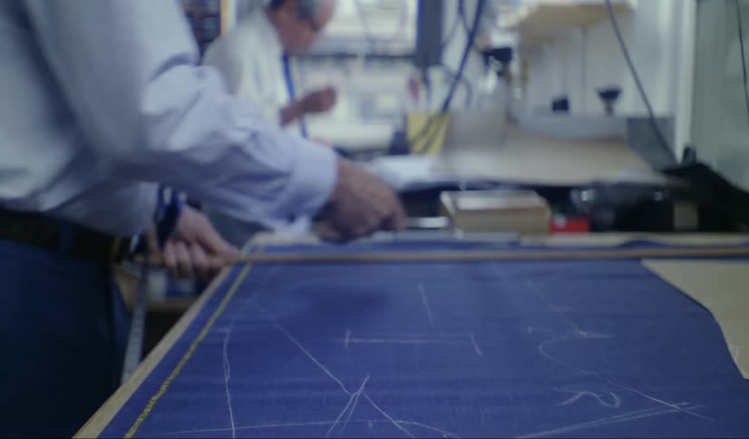 Close up of an older man using a ruler on blue paper and marking it with a white utensil while another man works near him