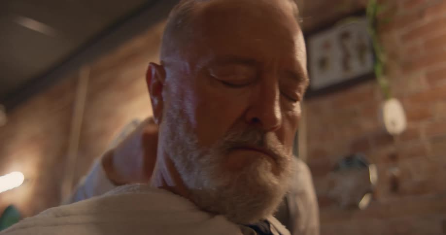 A man sits with his eyes closed as a barber wipes his head with a towel