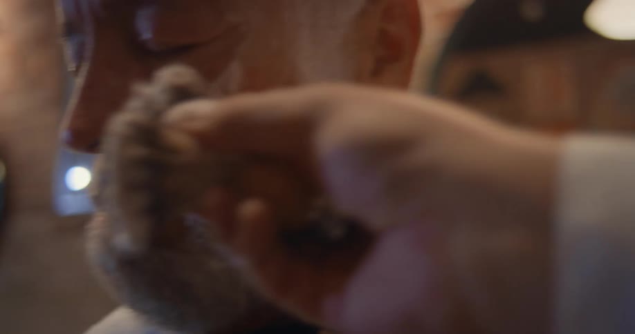 A clsoe up of a man getting shaving cream brushed on his head and face by a barber