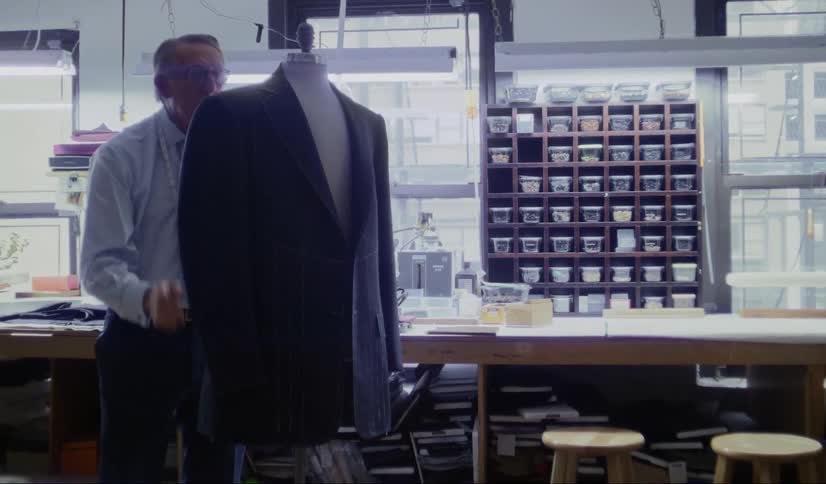 A tailor adjusts a suit jacket on a mannequin before walking out of frame