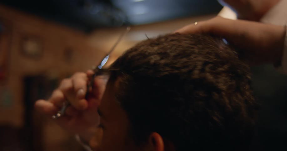 A barber combs and trims a young mans hair in a shop at night