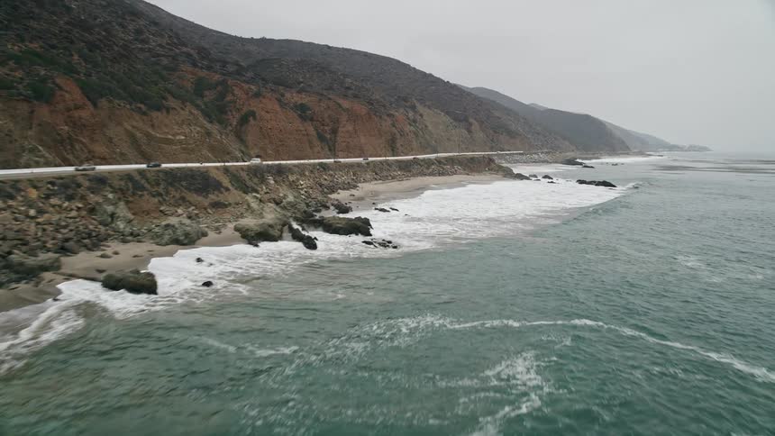 Low-flying 5K aerial footage captures waves crashing against the Malibu coast as traffic moves along Highway 1
