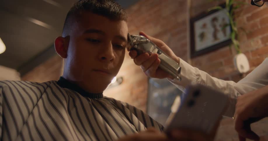 Man sitting in barber chair uses smartphone while the barber trims his hair