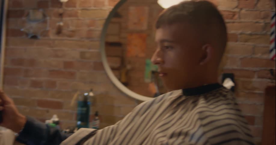 A teenager sitting at a barbershop holds a mirror to appreciate his haircut while swivelling his chair