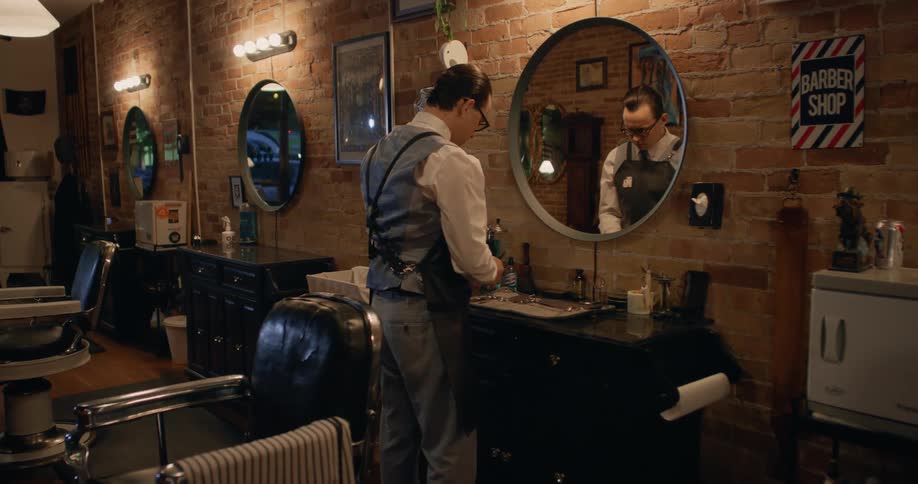 A man in a barber shop engaged in arranging his tools and standing in front of a mirror and a table