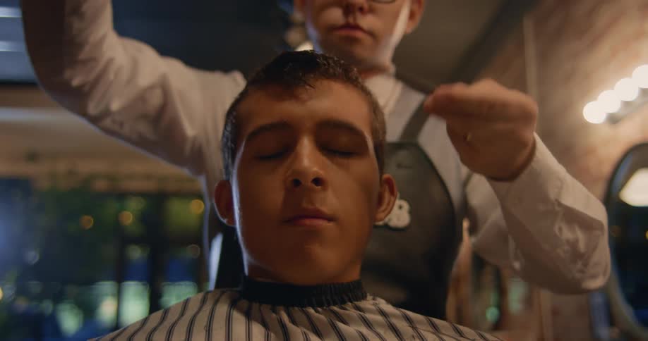 A close up of a young mans face with his eyes closed while he gets his hair cut by a barber using a comb and scissors