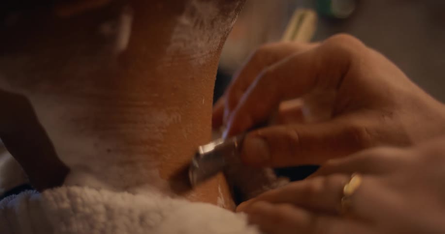 A barber is diligently working on his customers hair from the back of the neck using a sharp blade