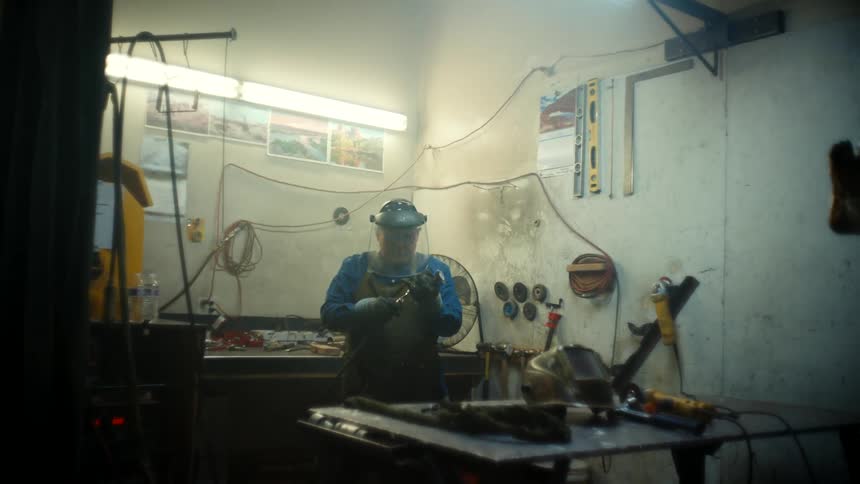 Man working in workshop connects cord to tool before approaching worktable across from sculpture