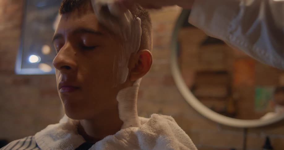 A young man sits in a barbers chair while a hand brushes shaving cream over his head and neck