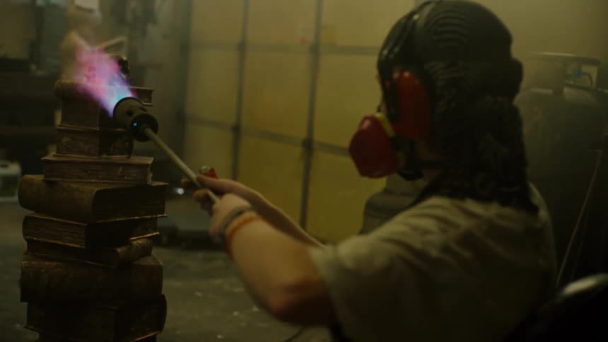 A person wearing a face covering is holding a blowtorch and scorching a stack of books rotating on a pedestal