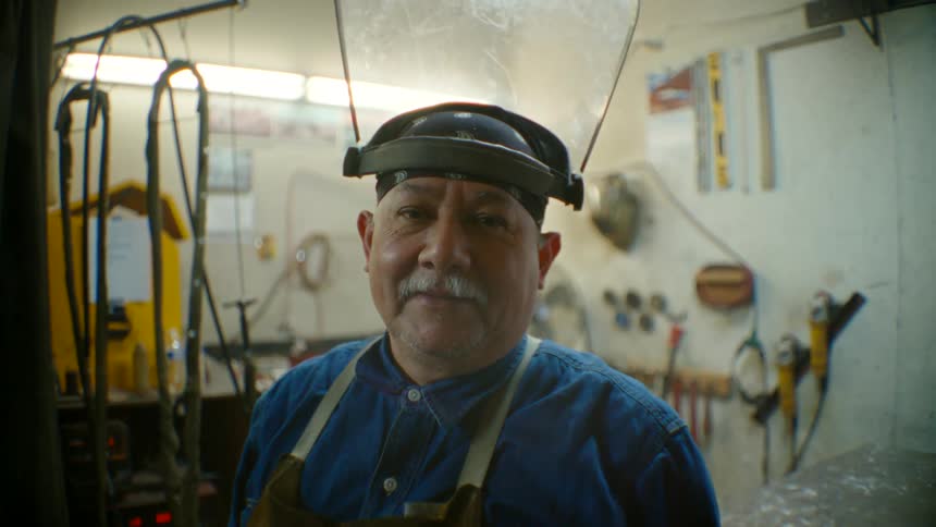 A portrait of a man wearing a work apron and face shield on his head while he stands in his work area