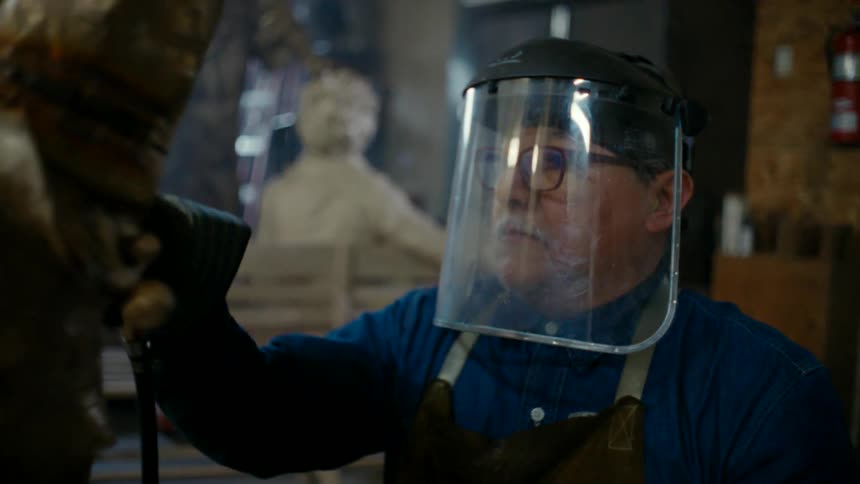 An elderly man uses a grinding tool in order to smooth a sculpture