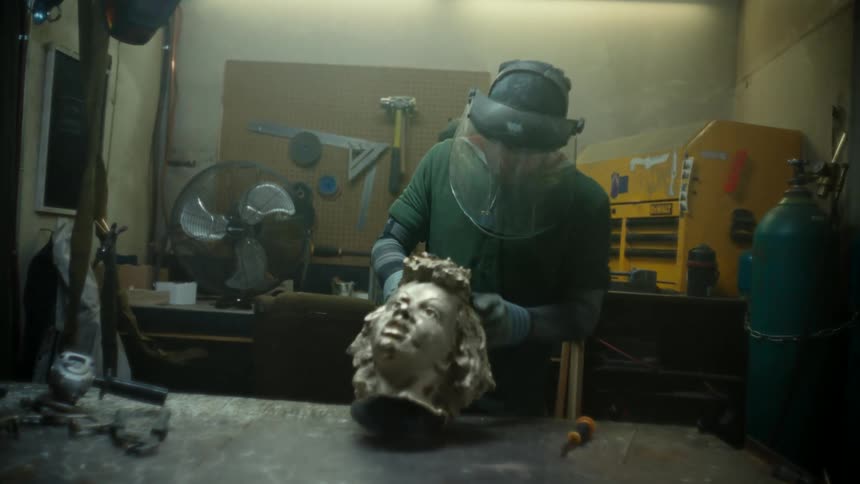 A man inside a room engaged in the art of molding a statue and puting on an overall and helmet