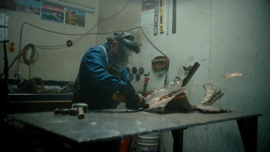 Older man wearing protective gear grinds metal sculpture pieces at workshop table