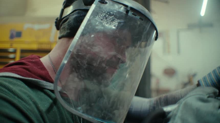 A man wearing a face shield and gloves working in a factory using a tool in his hands