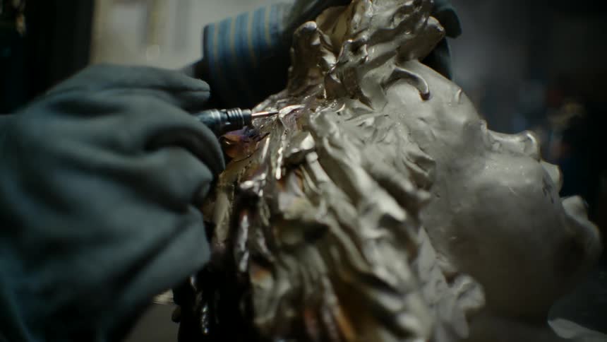 A sculptor is wearing gloves and holding a metal carving knife as they create a childs head from metal