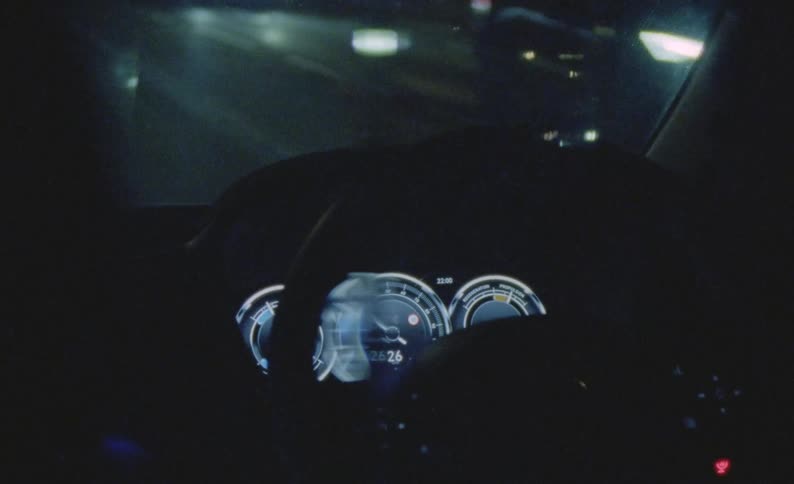 The drivers hands are on the steering wheel as the dashboard gauges glow in the darkness and they travel on a street at night