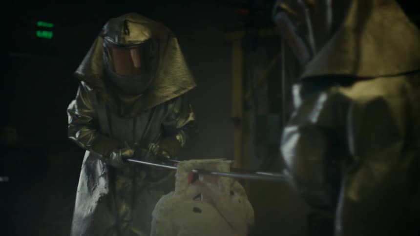 Two workers wearing a fire proximity suit carrying a hot crucible