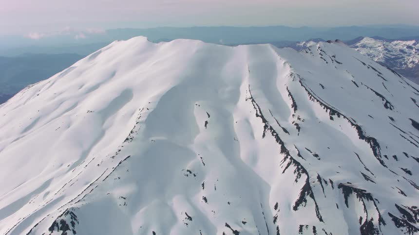 An aerial of a snow covered mountain or volcano in the middle of the day with mountains along the horizon