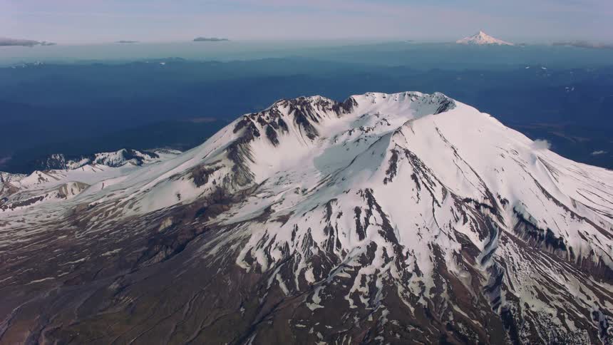 The sun shines upon a snow covered volcano and there are mountains and a snow covered peak in the distance