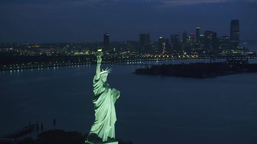 The statue of liberty is lit up at night across from downtown