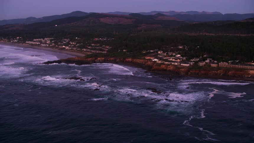 An aerial of a coastal village as waves crash against the beach at dusk with mountains in the distance