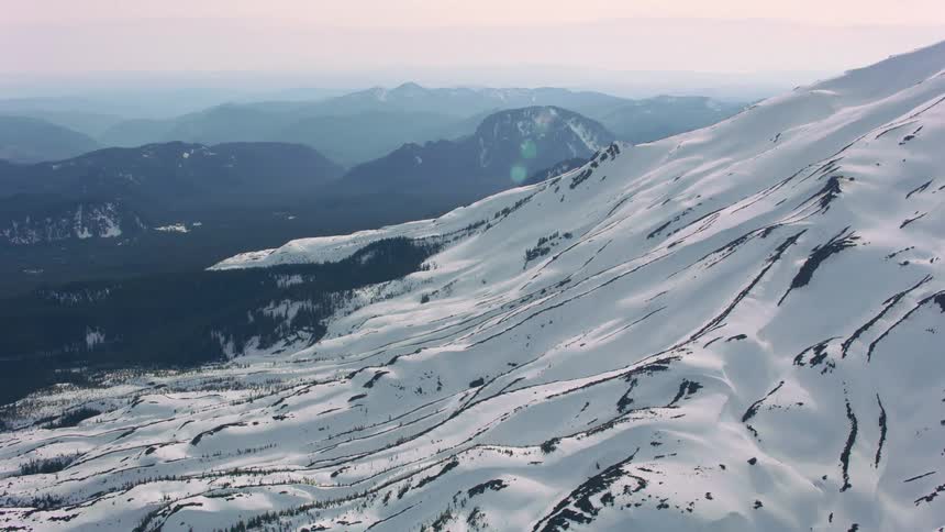 An aerial of snow covered mountain ridges with a mountainous landscape in the distance