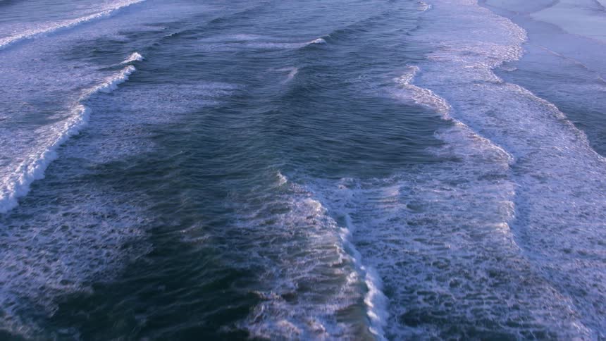 A birds eye view of the ocean waves rolling in toward the shore a crashing into flat ripples