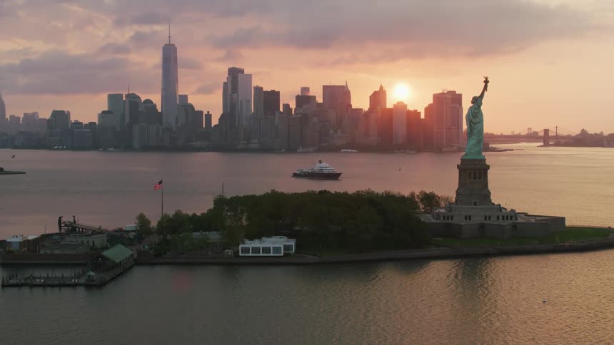 An aerial of the statue of liberty as the sun sets behind the city skyline