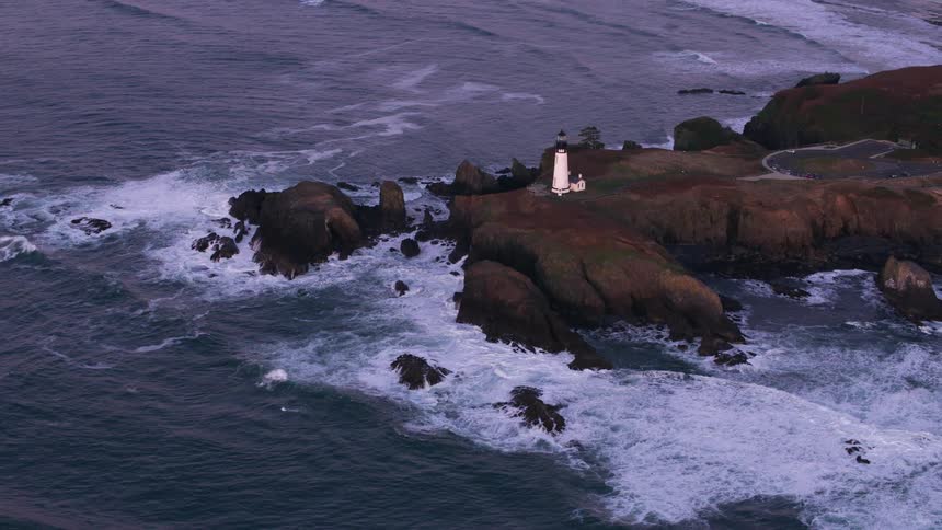 An aerial of a white lighthouse on the rock coastline as waves crash against the coast