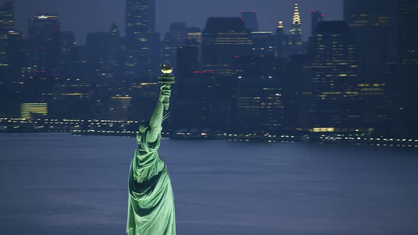 An aerial of the statue of liberty lit up at night with the silhouette of the city behind it