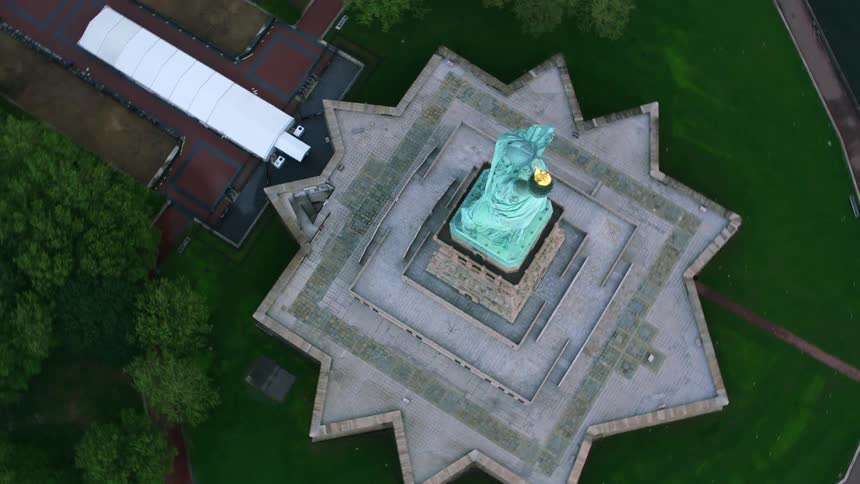 An aerial of the statue of liberty with lush green grass around it and the waterfront surrounding it