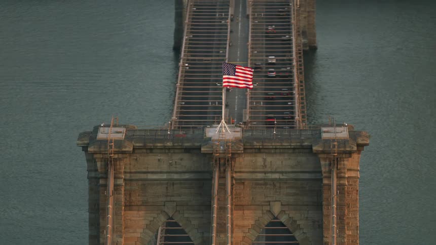 American flag flaps in wind on suspension bridge crossing water toward city