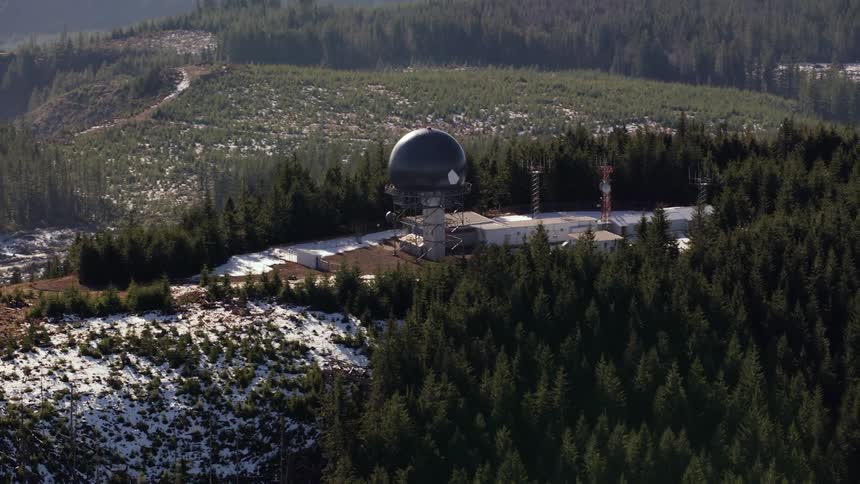 An aerial shot of an observatory building on a countryside hilltop with green trees and snow