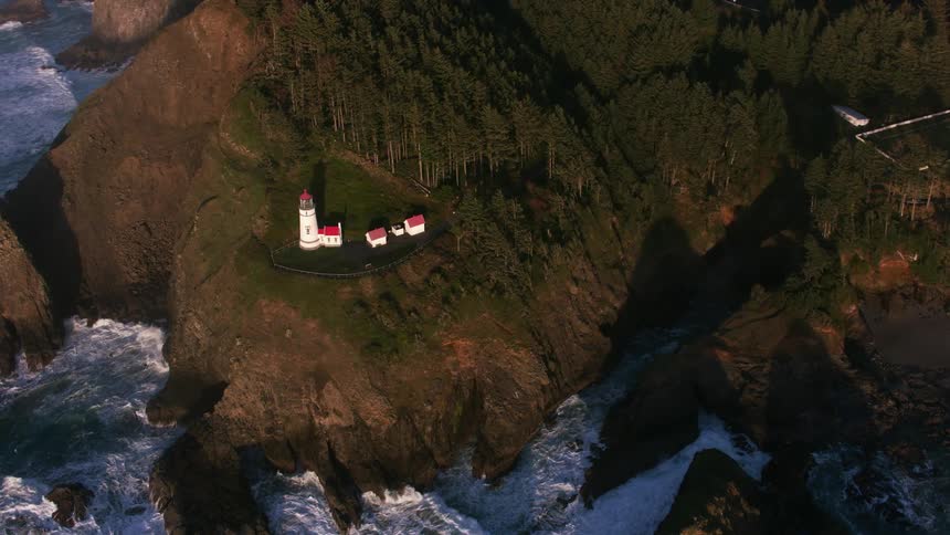 Aerial view of the ocean and a lighthouse at the coast on a forested rock at sunset
