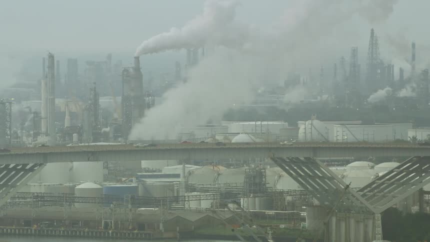 An aerial of traffic driving over a highway with a city in the background with smoke billowing out of the stacks