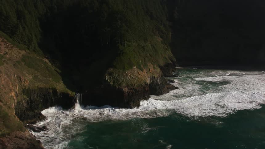 An aerial view of sea waves crashing against steep mossy cliffs