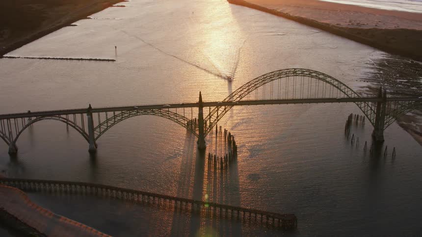 An aerial of an archway bridge near a beach with boats traveling on the water and cars on the bridge