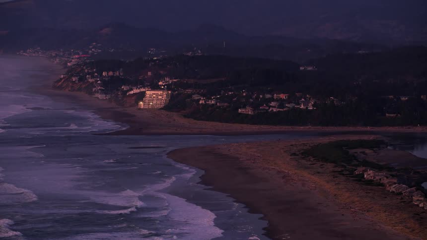 Waves roll into shore and over the beach as traffic moves on the streets of a coastal village with mountains in the distance